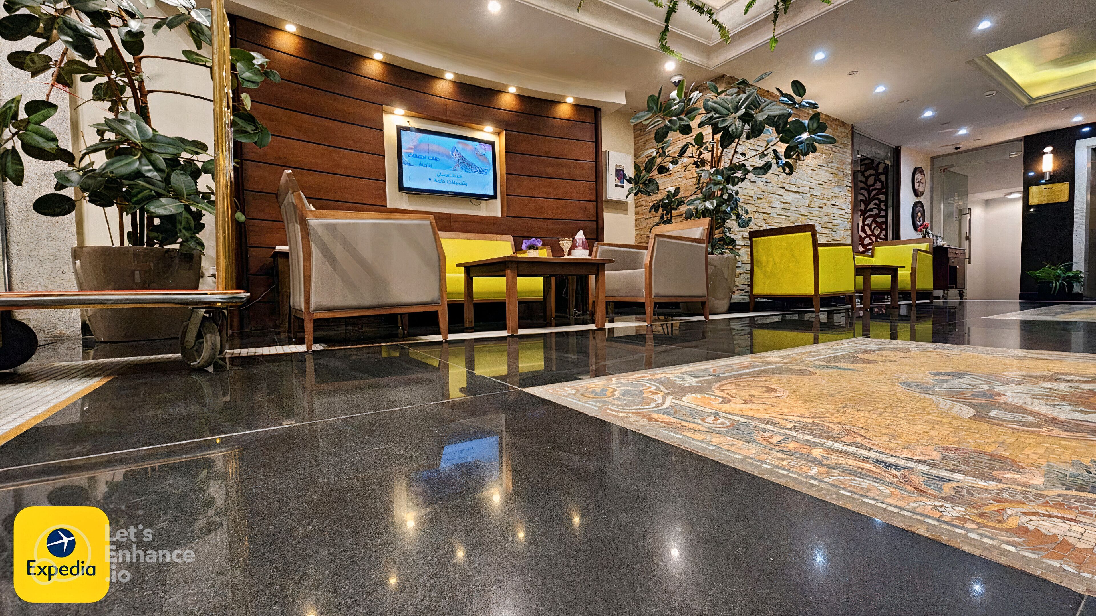 Eine Lobby mit einem Holzbüro, einer Sitzecke mit gelben Stühlen, einer großen Topfpflanze und einem Fernsehbildschirm.