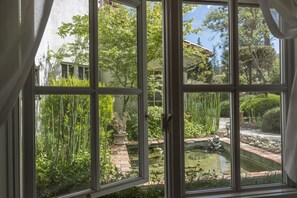 Vista pela janela de um jardim com lago, estátuas e vegetação exuberante.