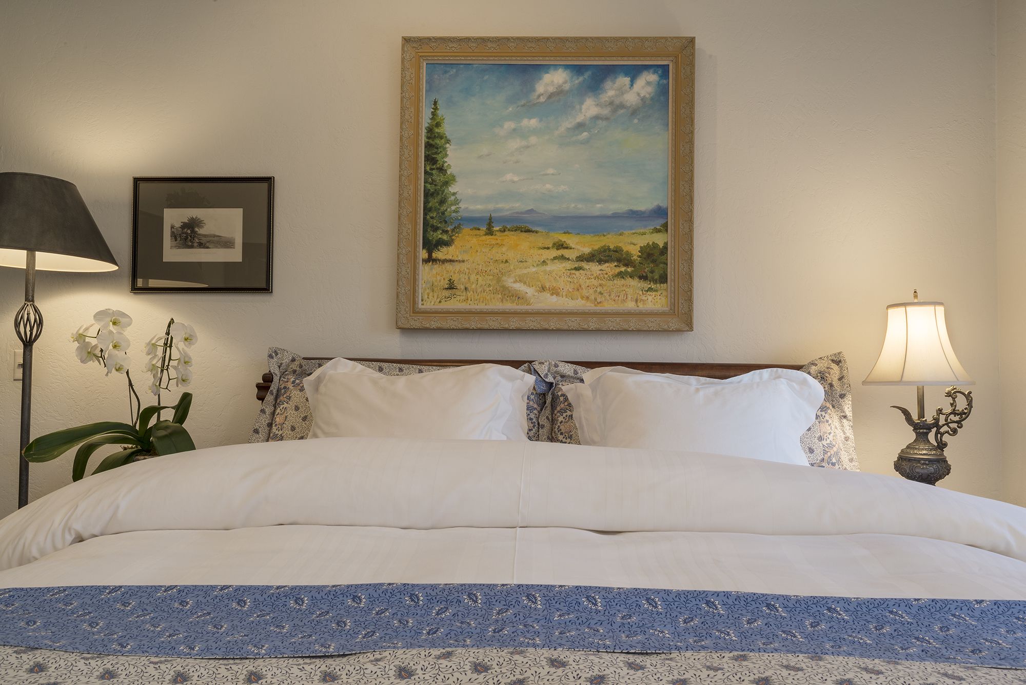 A neatly made bed with a blue and white patterned bedspread, a framed landscape painting above it, and two lamps on either side of the bed.