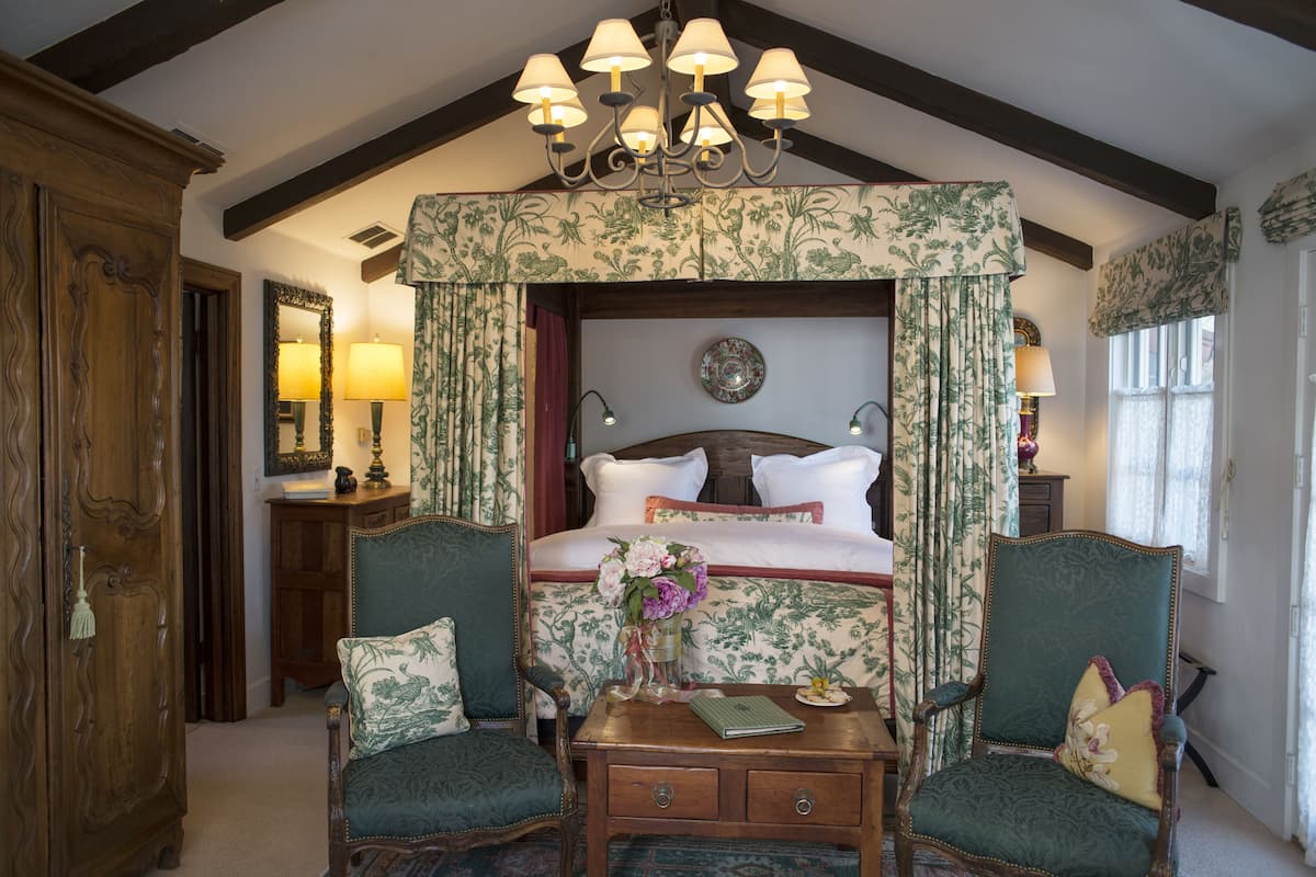 A bedroom with a four-poster bed, floral wallpaper, and a chandelier. There are two chairs and a nightstand with a vase of flowers.