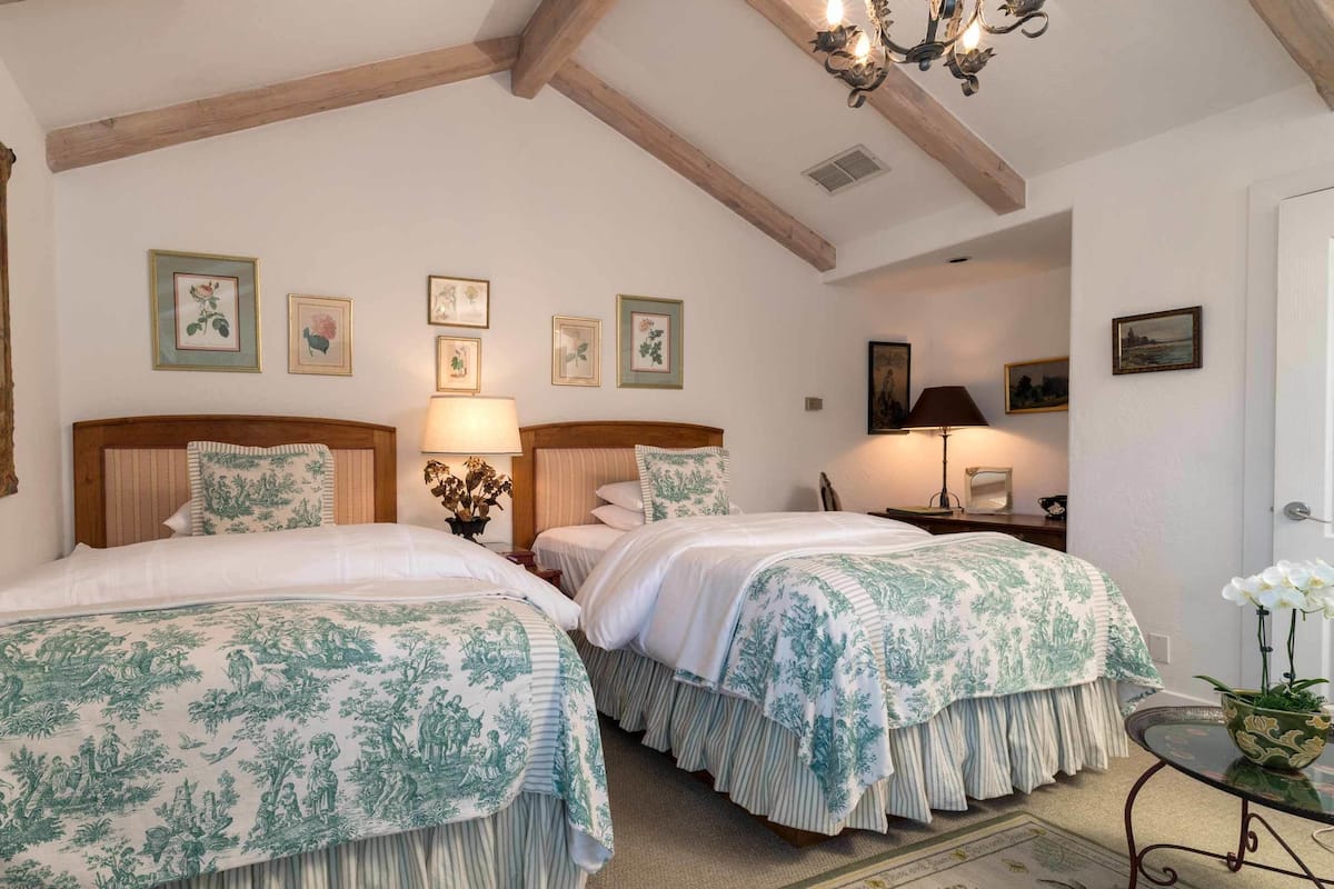 A bedroom with two beds, wooden headboards, a chandelier, and framed artwork on the walls.
