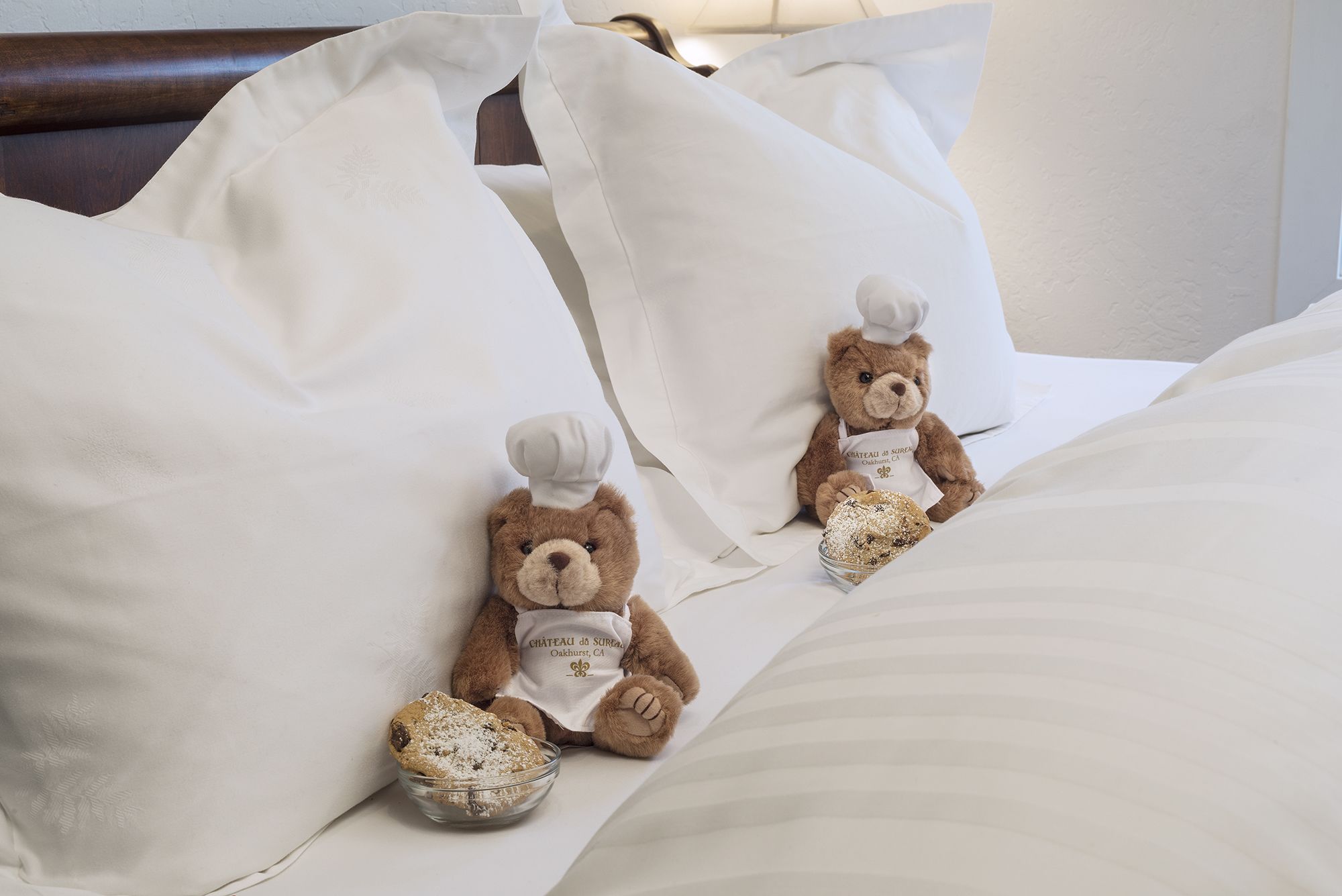 Two teddy bears on a bed with white bedding and pillows.