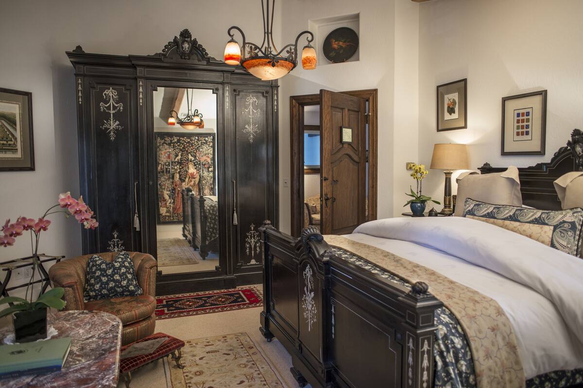 A bedroom with a large bed, a dark wooden wardrobe, a mirror, and a small table with a plant.