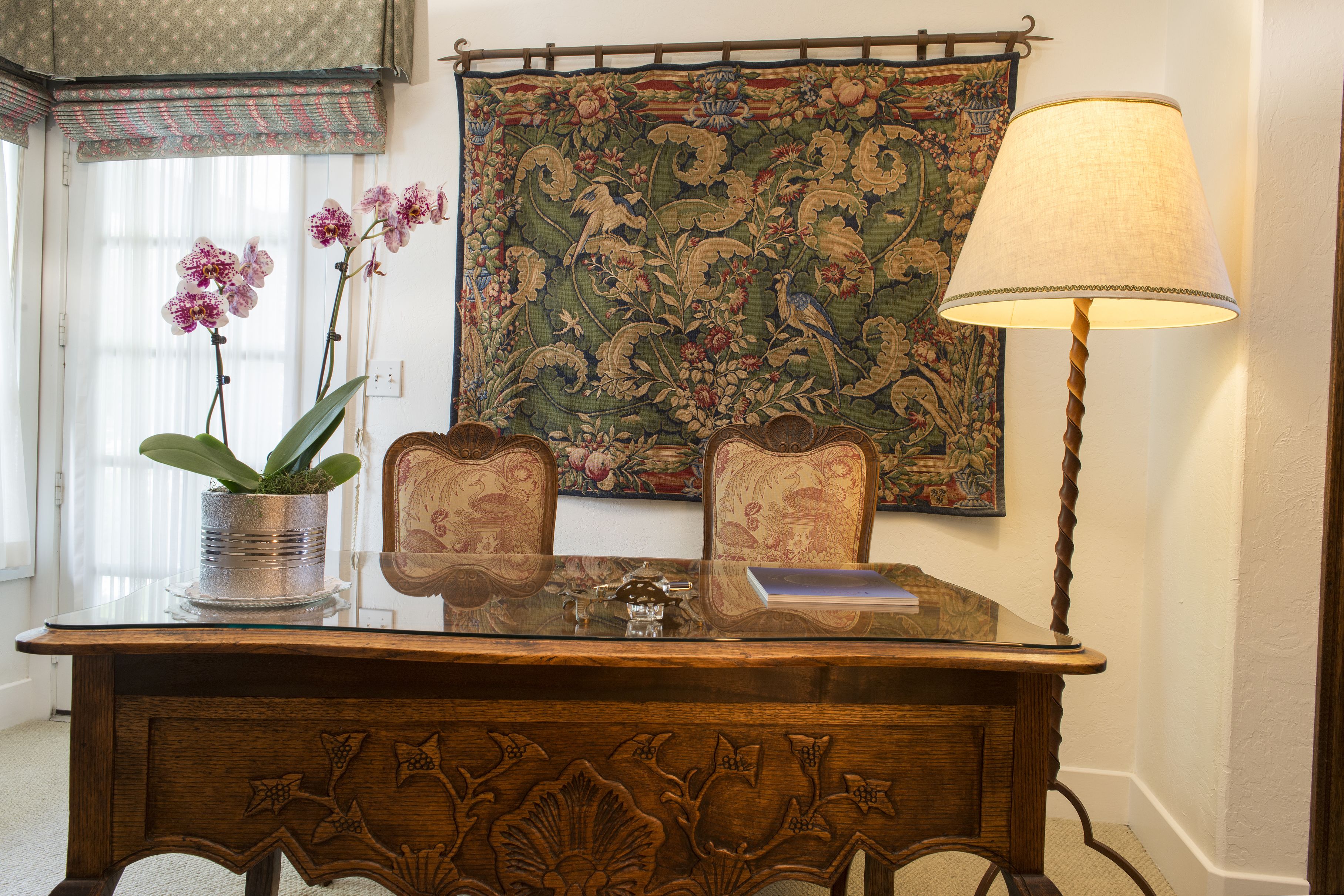 A wooden desk with a glass top, two chairs, a lamp, a potted plant, and a tapestry on the wall.
