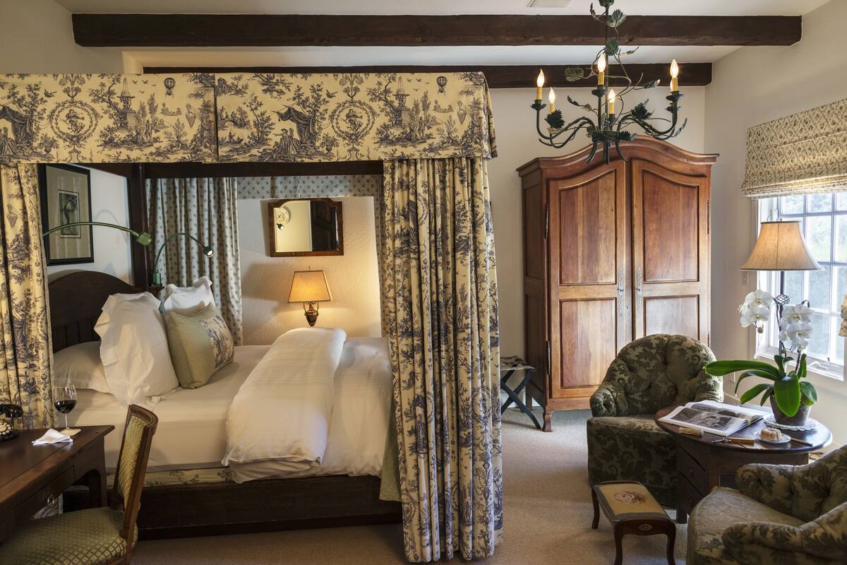 A bedroom with a canopy bed, a wooden wardrobe, a chair, a table with a lamp, and a window with curtains.