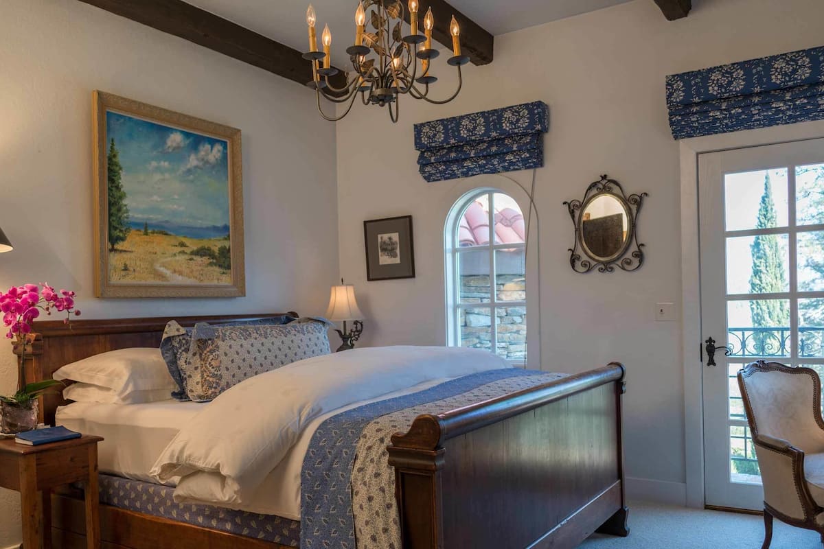 A bedroom with a bed, a nightstand, a lamp, a painting, a mirror, and a window with a view of trees.