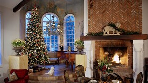 Une pièce chaleureuse, éclairée d’une lumière douce, où se trouve un grand sapin de Noël, une cheminée et un chien assis près du sapin.