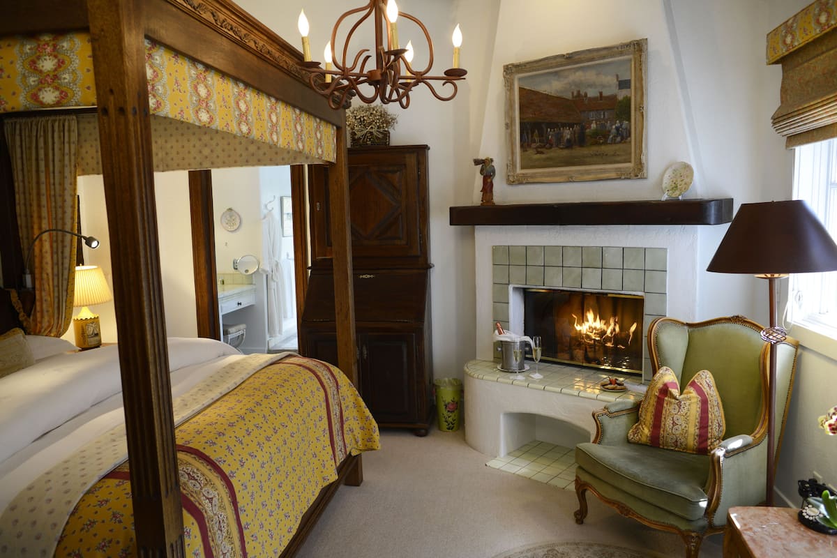 A traditional bedroom with a four-poster bed, a fireplace, a green armchair, and a desk with a lamp.