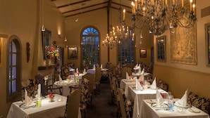 Une salle à manger élégante, avec plusieurs tables dressées pour le service, de somptueux lustres et de grandes fenêtres offrant une vue imprenable.