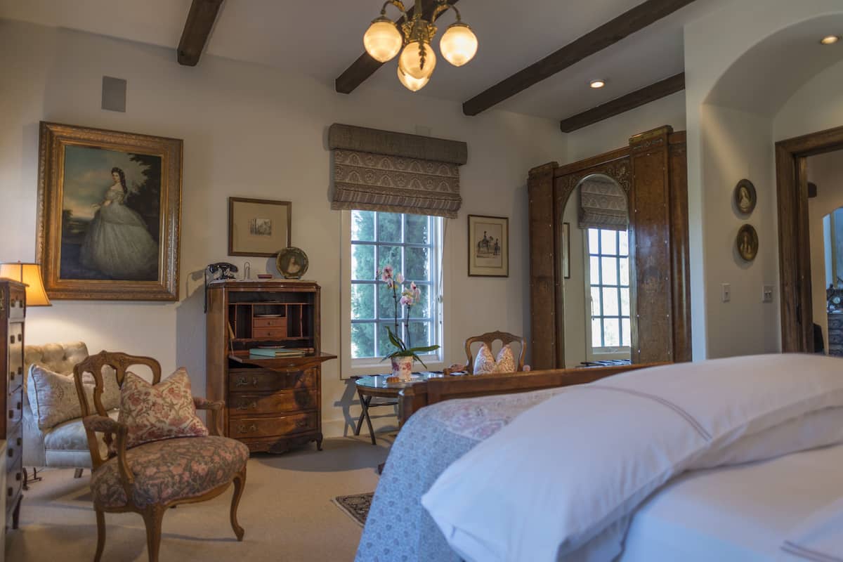 A bedroom with a large bed, a wooden dresser, a chair, and a desk. There is a window with a view of trees and a painting on the wall.