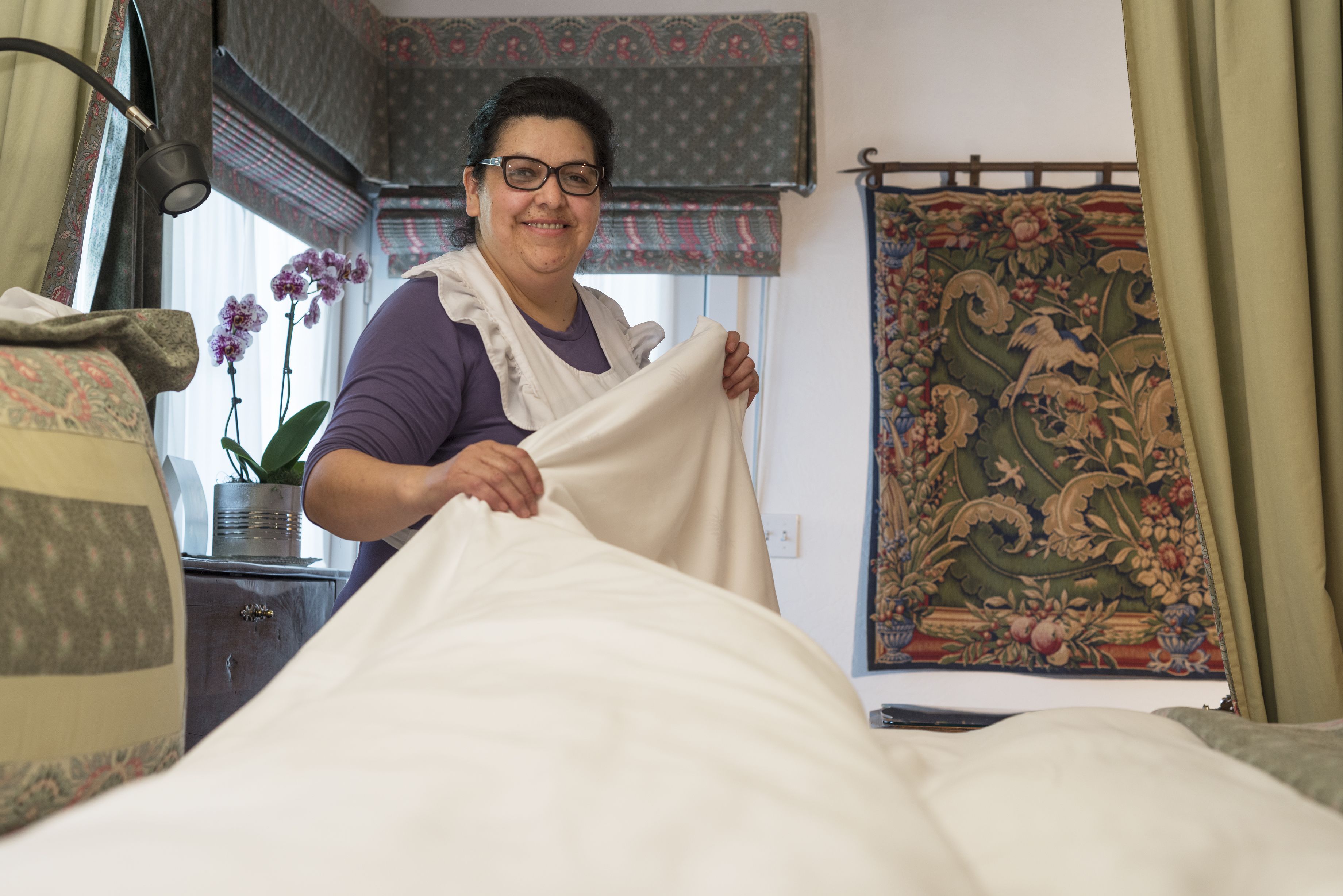 Una persona ajustando una cama en una habitación con un tapiz estampado, una planta en maceta y una ventana con cortinas.