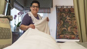 Una persona ajustando una cama en una habitación con un tapiz estampado, una planta en maceta y una ventana con cortinas.