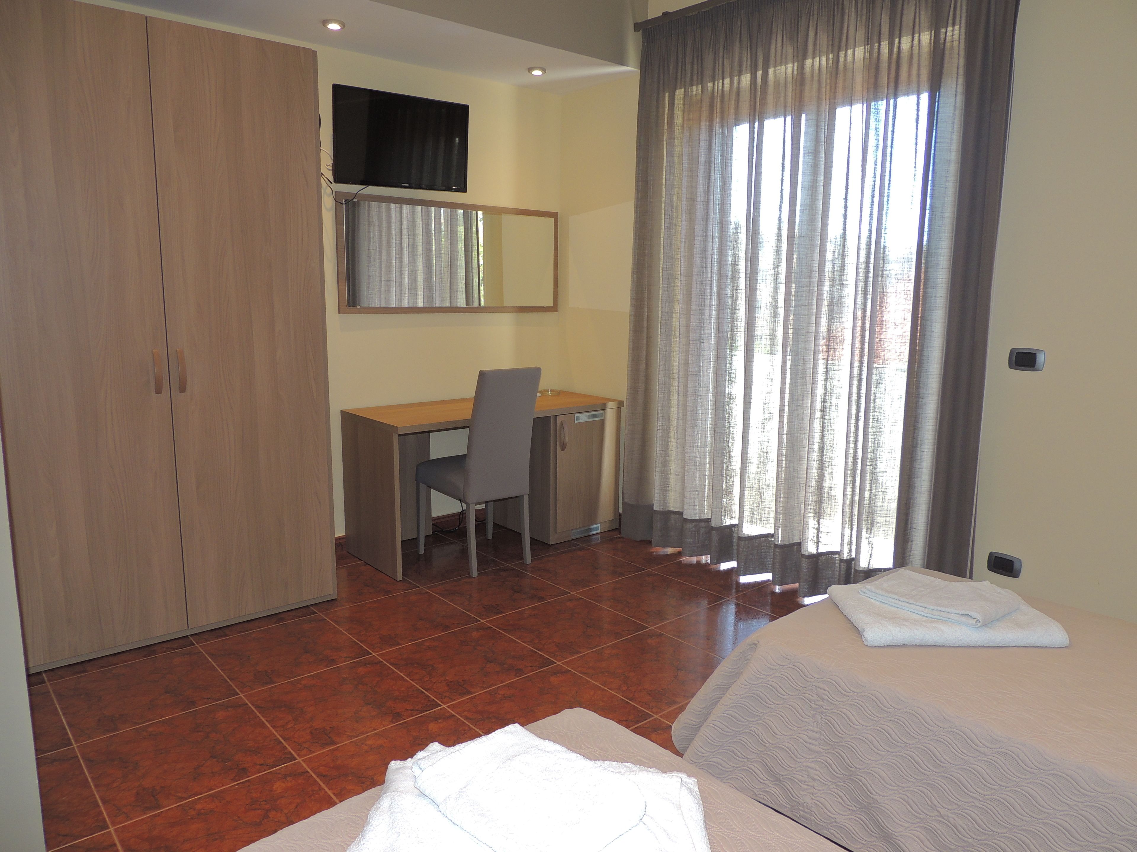 A hotel room with a wooden wardrobe, a desk with a chair, a bed with a white duvet, and a window with curtains.