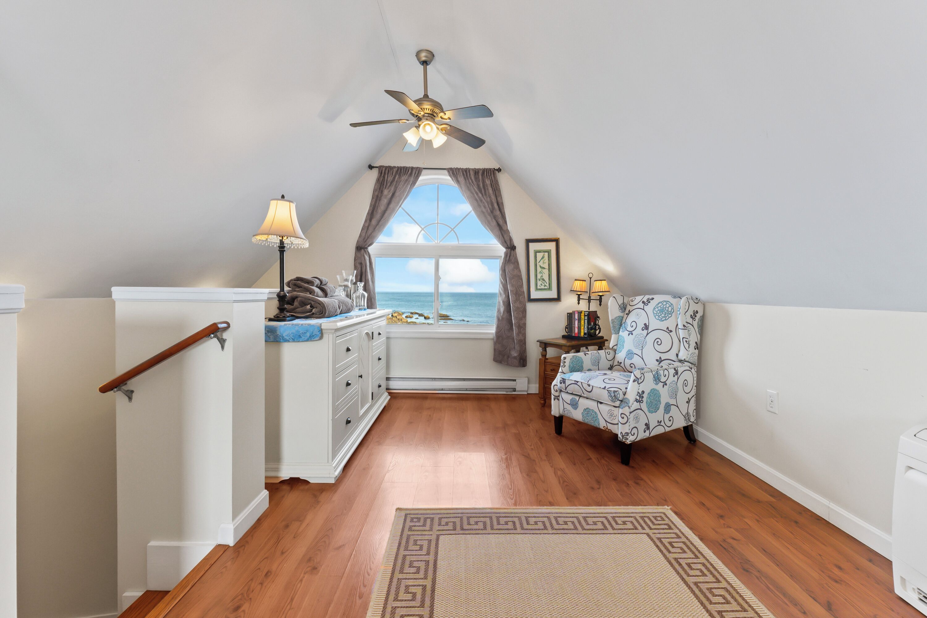 A cozy attic bedroom with a wooden floor, a ceiling fan, a window with curtains, a chair, and a small table with a lamp.