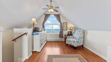 A cozy attic bedroom with a wooden floor, a ceiling fan, a window with curtains, a chair, and a small table with a lamp.