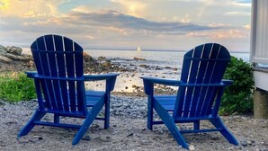 Zwei blaue Adirondack-Stühle, die auf einen felsigen Strand mit Blick auf das Meer und ein Segelboot im Hintergrund zeigen.