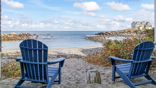 Zwei Adirondack-Stühle blicken auf eine felsige Küstenlinie mit Blick auf das Meer und ein Haus im Hintergrund.