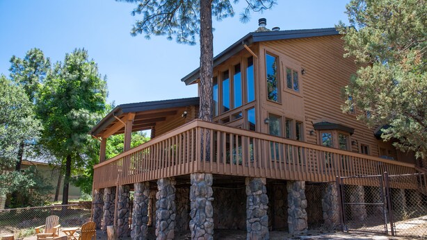 A log cabin with a stone foundation, a wooden deck, and a large window overlooking a forested area. - Cozy Mountain Log Cabin With Amazing Views!  (Pine)