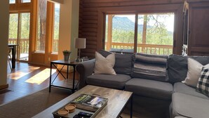 A cozy living room with a sofa, coffee table, and a view of a forest through large windows. - Cozy Mountain Log Cabin With Amazing Views!  (Pine)