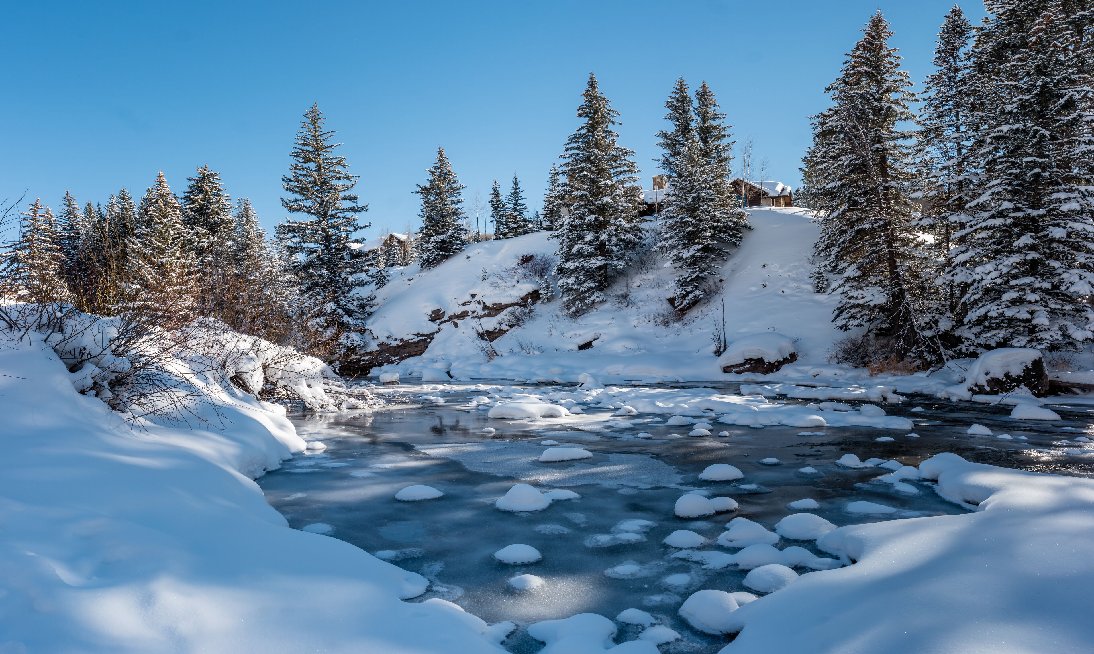 A snow-covered landscape with a frozen river and dense evergreen trees.