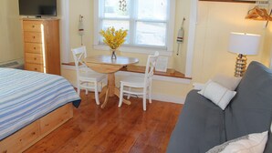 Ein Zimmer mit einem Bett, einem grauen Sofa, einer Holzkommode, einem Fernseher, einem Tisch mit Stühlen und einem Fenster mit Jalousien.