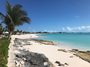 A beach with white sand, clear turquoise water, and a few people walking. There are palm trees and a small boat in the distance. - ROCK HILL: Modern Beach House with Spectacular Views & Resort Access (Stella Maris)