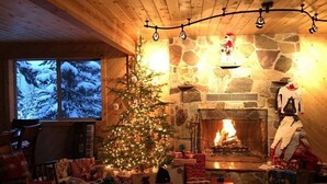 Una habitación acogedora con chimenea de piedra, un árbol de Navidad y un amplio ventanal con vistas nevadas.