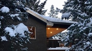 Una casa moderna de dos pisos con una terraza de madera y un ventanal grande. La nieve cubre el techo y los árboles.