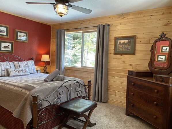 Un dormitorio con cama, una silla, cómoda, ventana con cortinas y una obra de arte en la pared.
