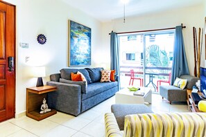 A living room with a sofa, armchair, and a coffee table. There is a door, a lamp, and a painting on the wall.