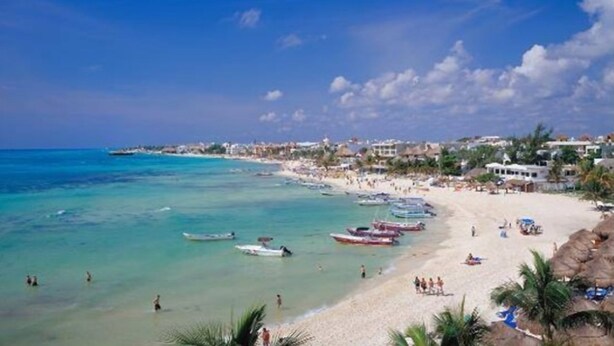 A beach with clear turquoise waters, several boats, and a sandy shore with people and umbrellas.