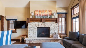 Un salon chaleureux avec une cheminée en pierre, des fauteuils confortables et une grande fenêtre agrémentée de rideaux.