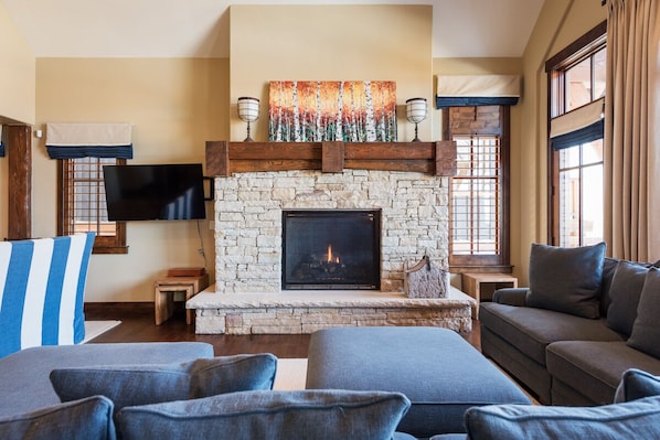 A cozy living room with a stone fireplace, comfortable seating, and a large window with curtains. - Last-Minute Savings! Cottage with Ski-in/Ski-out Access and Private Hot Tub! (Park City)