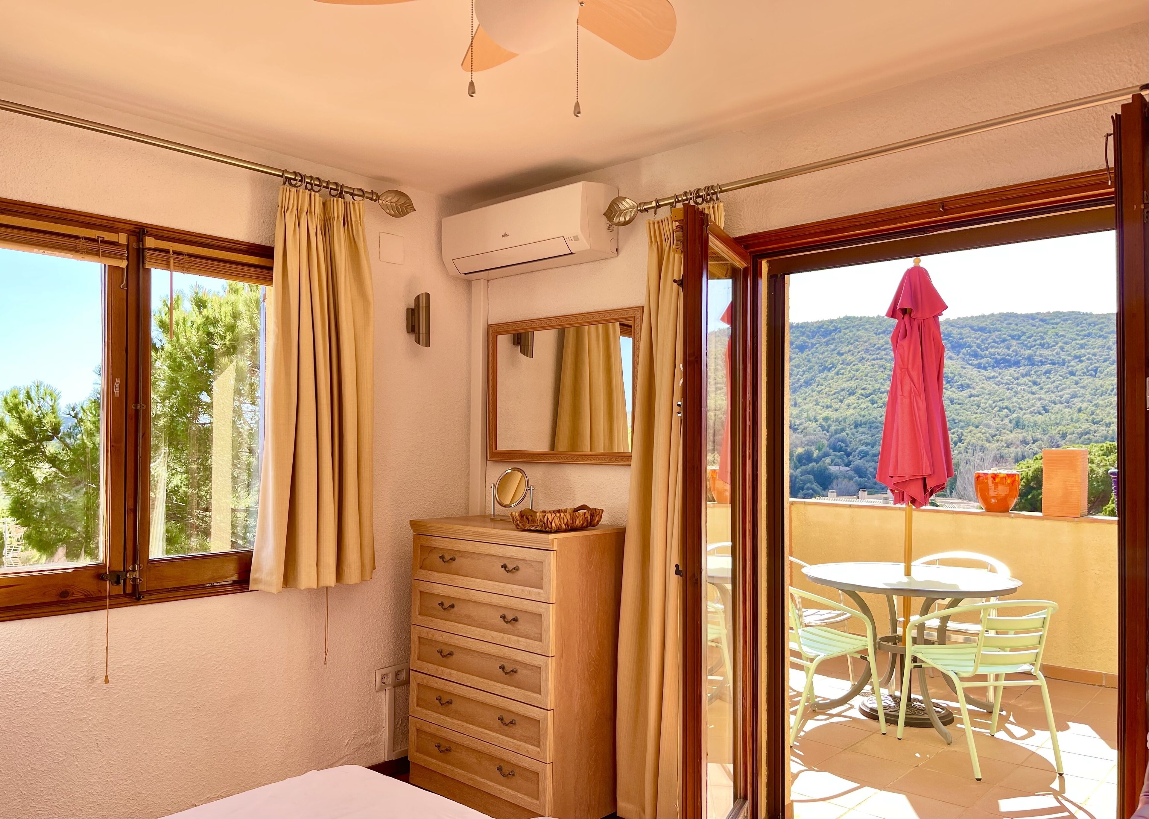 Ein Schlafzimmer mit einem hölzernen Schminktisch, einem Spiegel und einem Fenster mit Vorhängen. Es gibt einen Balkon mit Tisch und Stühlen sowie einen Blick auf einen bewaldeten Hügel.