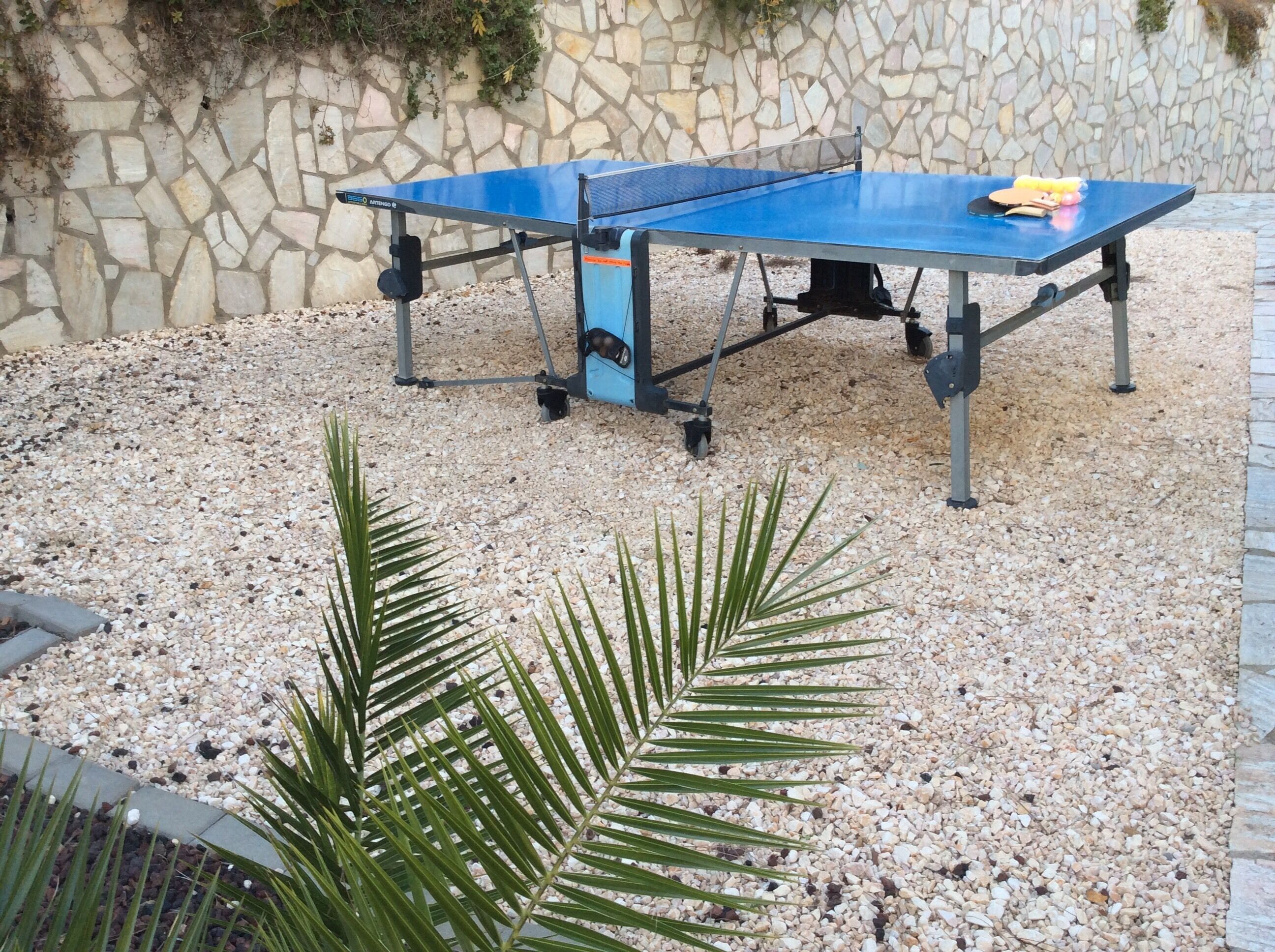 A ping pong table set up outdoors on a gravel surface with a stone wall in the background.