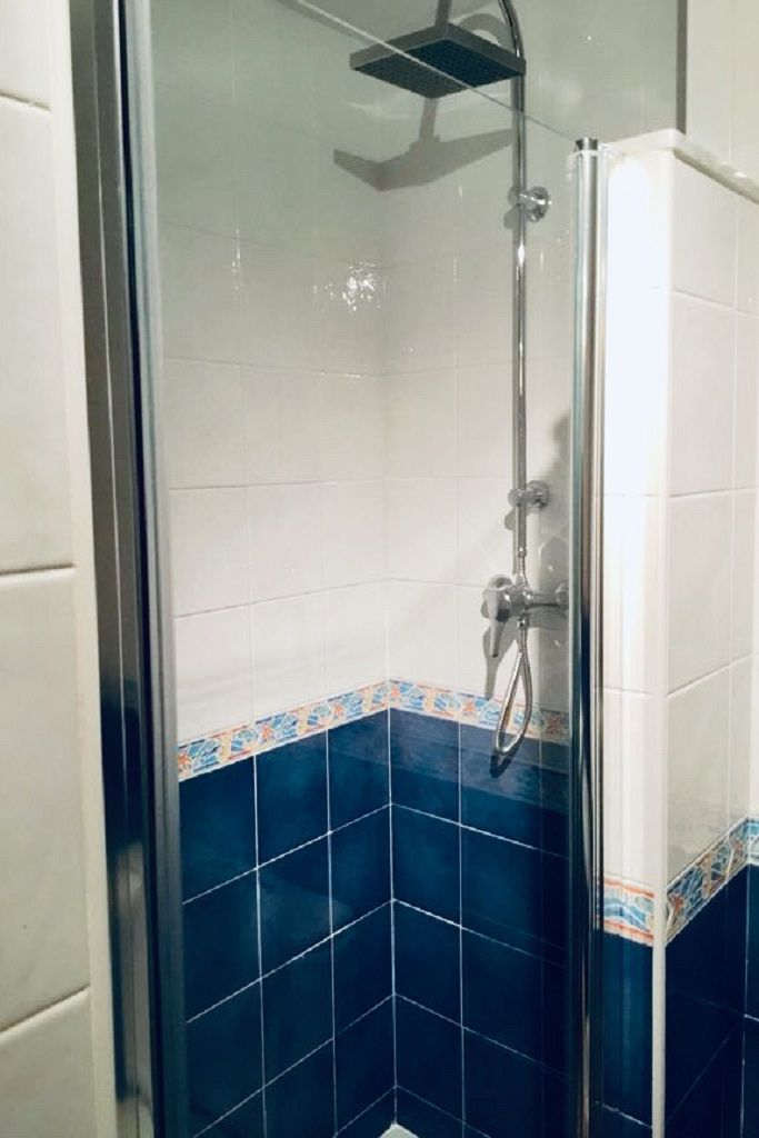 Ducha con mampara de vidrio, paredes de azulejos blancos y detalles azules en la parte inferior.