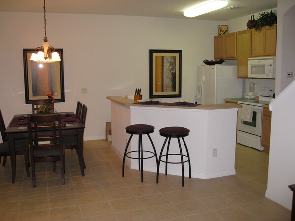 A kitchen with a dining area, including a table with chairs, a bar stool, a refrigerator, an oven, and a microwave.