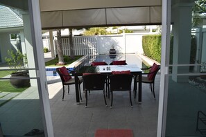 Un patio techado con una mesa y sillas de vidrio, una piscina y una valla blanca.