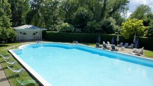 Une piscine rectangulaire à l’eau d’un bleu limpide, entourée d’une pelouse et d’une haie. Des chaises longues et des parasols sont disposés le long du bord de la piscine.