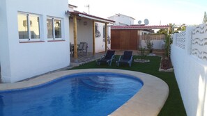A swimming pool with two lounge chairs, a small garden, and a white building with a tiled roof.