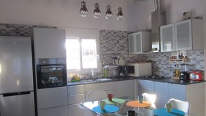Een moderne keuken met witte kasten, een koelkast, oven, magnetron en een spoelbak. Er is een eettafel met kleurrijke placemats en witte stoelen.