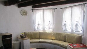 Een woonkamer met een gebogen bank, een klein tafeltje met een rode tafelkleed, een klok aan de muur en twee ramen met transparante gordijnen.