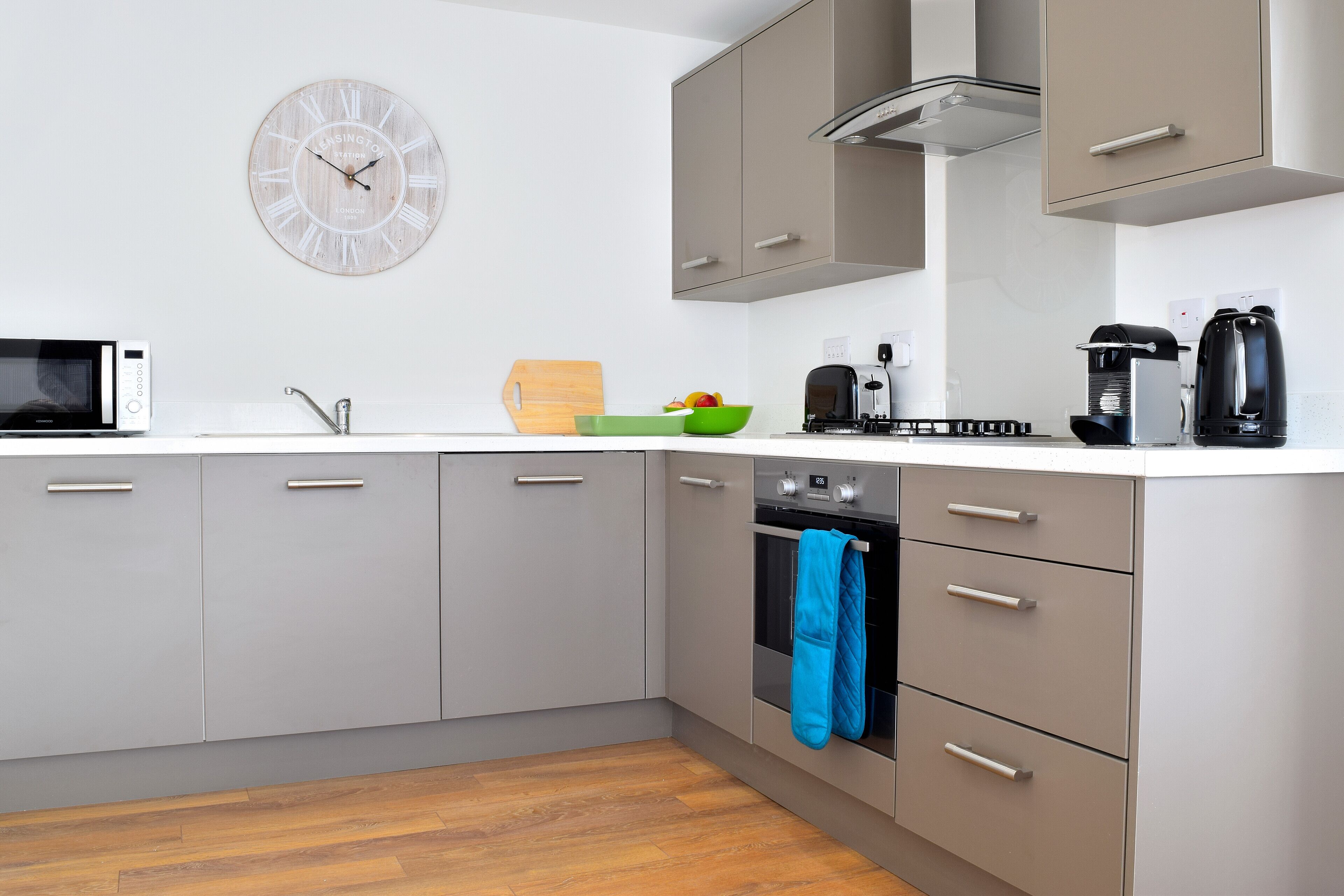 A modern kitchen with grey cabinets, a microwave, a kettle, and a clock on the wall.