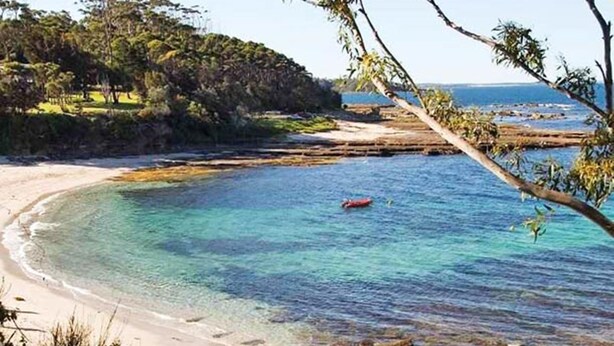 Una playa costera con agua turquesa cristalina, costa rocosa y una vista distante de una zona boscosa.