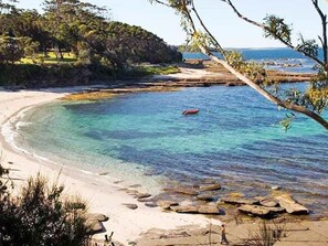A coastal beach with clear turquoise water, rocky shoreline, and a distant view of a forested area.