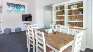 Un comedor con una mesa de madera y sillas blancas, un televisor en la pared y una estantería con cristalería.
