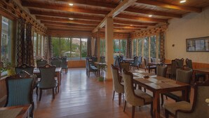 Un'ampia sala da pranzo con tavoli e sedie in legno, grandi finestre con tende e vista sul verde.