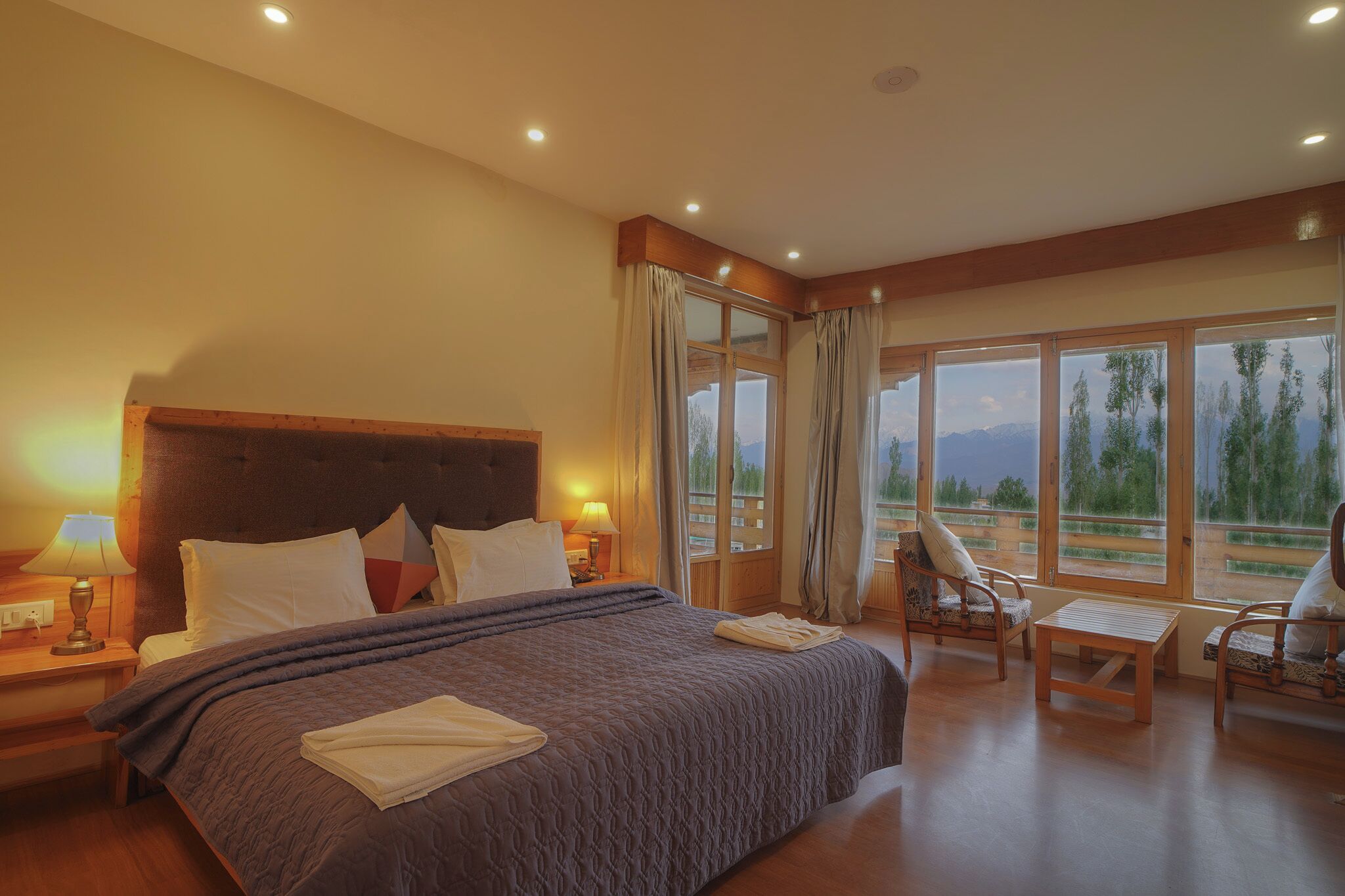 Ein Hotelzimmer mit einem großen Bett, einem hölzernen Kopfteil, einem Nachttisch mit Lampe, einem kleinen Tisch, zwei Stühlen und Blick ins Freie durch große Fenster.