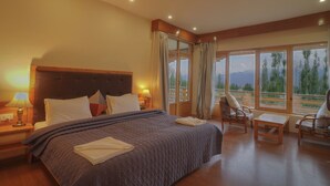 Camera d'albergo con un letto grande, una testiera in legno, un comodino con una lampada, un tavolino, due sedie e vista sull'esterno attraverso ampie finestre.