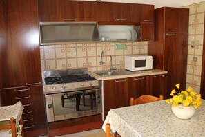 A compact kitchen with wooden cabinets, a stove, oven, microwave, and a small table with a vase of flowers. - "My Home", 2 rooms, not far from the beaches and in the historic center of Marsala (Marsala)